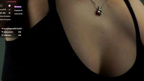 Media: A close-up photograph of a woman's cleavage in a low-cut black top, featuring a silver necklace and a small mole on her right breast. The background is blurred, focusing on her chest area.