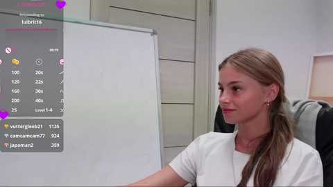 Media: A photograph of a young woman with long, straight brown hair, wearing a white shirt, sitting in a chair, in an office setting. A virtual reality headset is on her head, displaying a screen with social media stats.