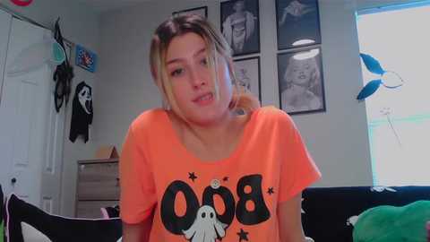 Media: Video of a young woman with light skin and blonde hair in a room with gray walls, wearing an orange \"Ghostbusters\" t-shirt. Background includes framed photos, a stuffed toy, and Halloween decorations.