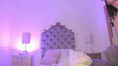 Media: Video of a modern bedroom with purple lighting, featuring a tufted headboard, a white lamp, and a white bed with white linens.