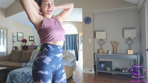 Media: Video of a fit woman in a purple tank top and blue tie-dye leggings stretching in a modern living room with wooden floors, a couch, and abstract art.