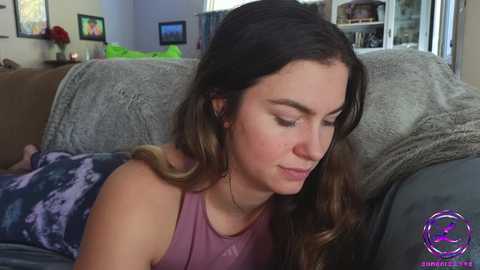 Media: Video of a young woman with long brown hair, wearing a purple tank top, sitting on a gray couch in a living room with a blue blanket.