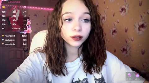 Media: Video of a young woman with wavy brown hair, wearing a white shirt, seated in a room with floral wallpaper and a door visible in the background.