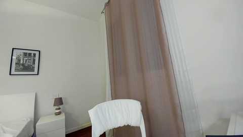 Media: Video of a minimalist bedroom with beige curtains, white chair, white bed, and a black-and-white framed photo on a white nightstand.