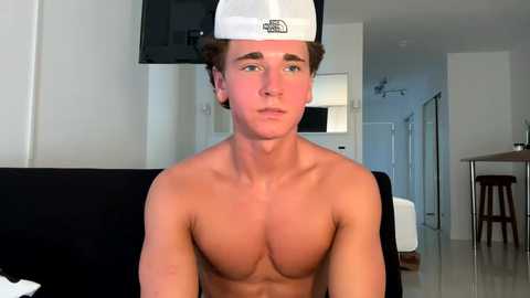 Media: Video of a shirtless young man with a white cap, sitting on a black couch in a modern, minimalistic living room with a TV, white walls, and a wooden dining table in the background.