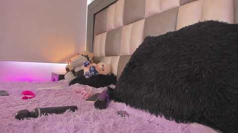 Media: Video of a plush toy bear lying on a fluffy, light purple carpet, surrounded by scattered sex toys, including a red vibrator, in a modern, dimly lit bedroom with a padded headboard.