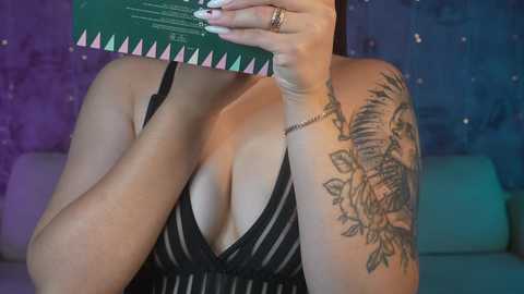 Media: Video of a woman with light skin, wearing a black, low-cut, pinstriped dress, covering her face with a book. She has a large floral tattoo on her left shoulder and is seated against a purple and blue backdrop.