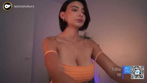 Media: Video of a young woman with short dark hair, wearing an orange off-shoulder top, standing in a dimly lit room.