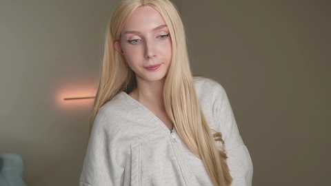 Media: Video of a young, fair-skinned woman with long, straight blonde hair, wearing a light-colored, loose-fitting top, standing against a plain, beige wall with a dimly lit light fixture in the background.