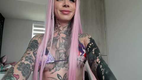 Media: Video of a tattooed woman with long pink hair, wearing a purple bikini, standing indoors with white walls and a window in the background.