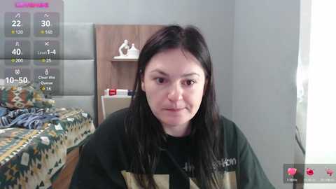 Media: Video of a young woman with long black hair, fair skin, and light makeup, wearing a black graphic t-shirt, sitting on a bed with a patterned blanket. Background shows a wooden headboard and a nightstand with a white vase.