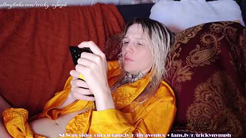 Media: Video of a fair-skinned, blonde woman with wet hair, wearing a yellow robe, holding a black phone, lying on a red and gold patterned cushioned sofa.