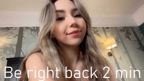 Media: Video of a young woman with long, wavy blonde hair and light skin, smiling slightly, captioned \"Be right back 2 min.\" in white text. Background features a softly lit room with green and grey floral wallpaper.