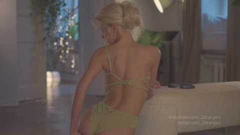 Media: Video of a slender, blonde woman with fair skin, wearing a lime green lingerie set, leaning against a massage table in a softly lit, modern indoor setting.