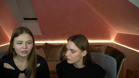 Media: Video of two young women with light skin, long brown hair, and black outfits, seated on chairs in a modern, dimly lit room with a sloped ceiling and soft pink walls.