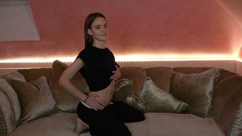 Media: Video of a young woman with fair skin, brown hair, and small breasts, wearing a black crop top and black pants, kneeling on a beige couch, touching her pregnant belly.