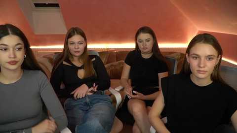 Media: Video of four young women with light skin, long straight hair, and dark clothes seated on a plush, orange sofa in a modern, dimly lit room with warm lighting.