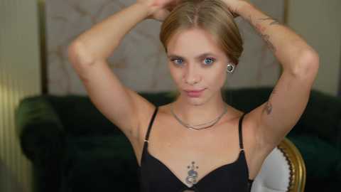 Media: Video of a young woman with blonde hair, blue eyes, and fair skin, wearing a black tank top, silver necklace, and ear gauges. She's adjusting her hair, revealing tattoos on her arms. Background shows a green couch and abstract artwork.