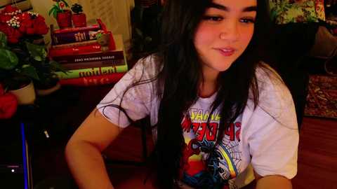 Media: Video of a young woman with long black hair, fair skin, and a small nose, wearing a white t-shirt with a colorful graphic, sitting indoors with books and plants in the background.