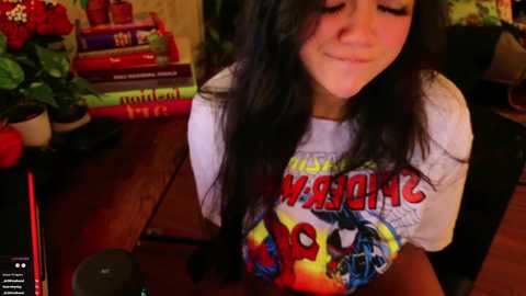 Media: Video of a young woman with long black hair, wearing a white \"Harry Potter\" T-shirt, smiling, seated at a wooden table with books and plants in the background.