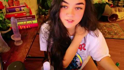 Media: Video of a young woman with long, wavy dark hair, wearing a white T-shirt with colorful graphic, sitting on a wooden floor amidst books, potted plants, and a purple dildo.