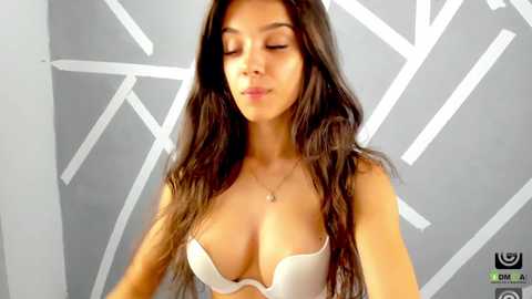 Media: Video of a young woman with long, dark hair, wearing a white bra, standing against a geometric grey and white wall.