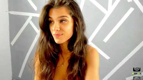 Media: Video of a young woman with long, wavy brown hair, fair skin, and a neutral expression, standing against a grey wall with white geometric patterns. She's topless.