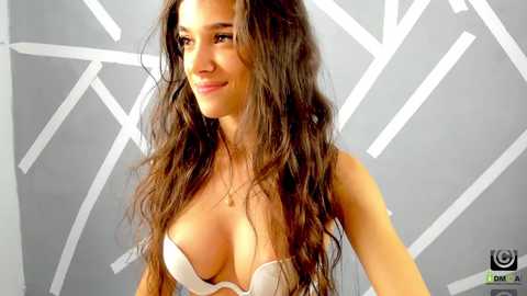 Media: Video of a smiling woman with long, wavy brown hair, wearing a light pink bra with lace detailing, against a modern, geometrically patterned grey wall.