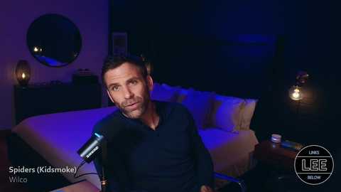 Media: Video of a man with short brown hair and a beard, wearing a dark shirt, seated in a dimly lit bedroom with a bed, nightstand, and lamp.