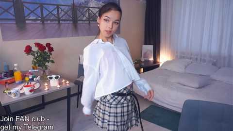 Media: A young woman with a light complexion, dark hair, and a slender figure stands in a cozy bedroom, wearing a white blouse and plaid skirt, surrounded by flowers and candles.