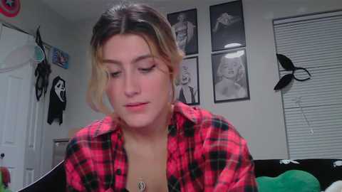 Media: Video of a young woman with fair skin, blonde hair, and freckles, wearing a red and black plaid shirt, looking contemplative in a bedroom with posters and a door.