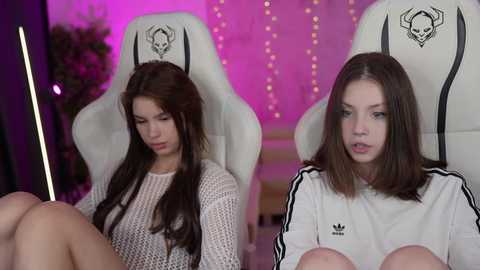 Media: Video of two young women, one with long brown hair, wearing a white Adidas hoodie, and the other with straight hair, in white chairs, in a pink-lit room.