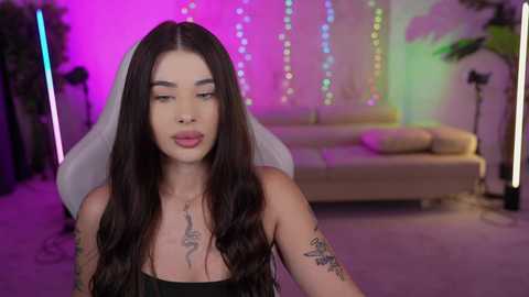 Media: A video of a tattooed woman with long dark hair in a purple-lit room, wearing a black strapless top.