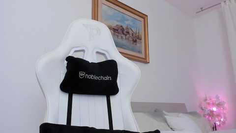 Media: Video of a white gaming chair with a black \"robochair\" cushion on it, placed in a minimalist room with a framed painting on the wall and a pink lamp in the background.