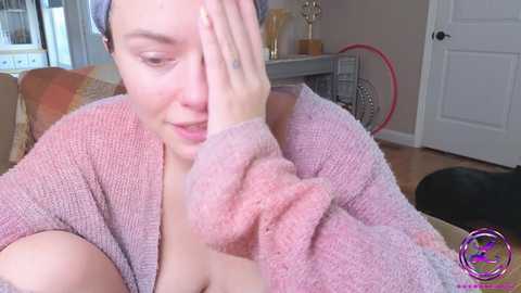 Media: A video shows a light-skinned woman with short hair, wearing a pink cardigan, covering her face with her hand, seated on a beige couch in a cozy room with wooden floors, a white door, and a fan.