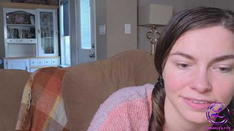 Media: Video of a smiling young woman with light skin and brown hair in a braided ponytail, sitting on a beige couch with a plaid blanket. The background features a glass cabinet and door, and a lamp.