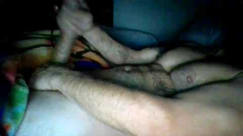 Media: Video of a close-up view of a man's legs and genitals in a dimly lit room. His legs are intertwined with a green and blue striped blanket. The image is grainy and somewhat blurry, with a dark background.