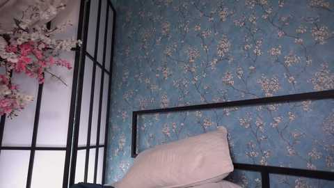 Media: Video of a cozy bedroom corner featuring a light blue, floral-patterned wall, a black folding screen with pink cherry blossoms, and a beige pillow on a white bed.