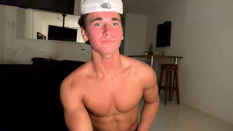 Media: Video of a shirtless young man with a white chef's hat, standing in a dimly-lit kitchen. He has a toned, muscular physique, light skin, and short brown hair. The background shows a black stove, a microwave, and a wooden bar counter with stools.