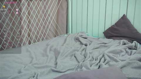 Media: Video of a messy bed with gray blanket, pillows, and a white metal headboard with a diamond pattern. Background includes vertical green blinds and a pink toy car.