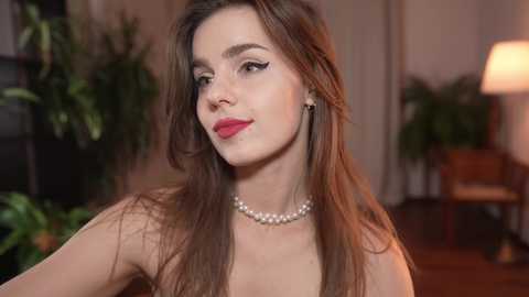 Media: Video of a young woman with fair skin and long brown hair, wearing red lipstick, a pearl necklace, and a strapless top, set against a softly lit, indoor background with potted plants.