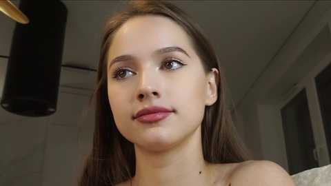 Media: Video of a young Caucasian woman with light skin, straight brown hair, and fair complexion, wearing a light-colored top, looking contemplative, set in a modern, dimly-lit room with a black cylindrical lamp.