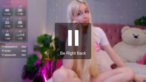 Media: Video of a blonde woman in white lingerie, sitting on a bed with a teddy bear, surrounded by plants, with a digital clock and \"Be Right Back\" text overlay.