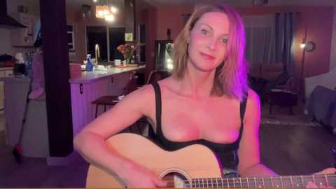 Media: Video of a smiling woman with shoulder-length blonde hair, wearing a black sleeveless top, playing an acoustic guitar in a dimly lit living room with purple lighting.