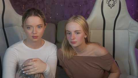 Media: Video of two young women with fair skin, one with blonde hair and the other with brown hair, sitting in white gaming chairs against a purple backdrop.