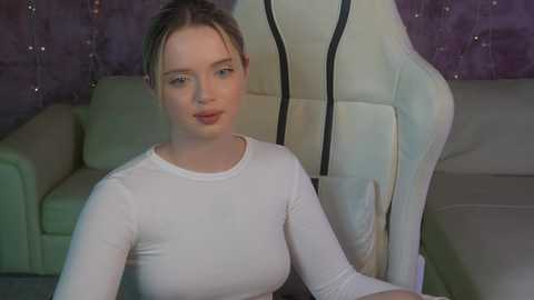 Media: Video of a young Caucasian woman with fair skin, light brown hair tied back, wearing a white long-sleeved top, sitting on a beige couch in a room with purple walls.