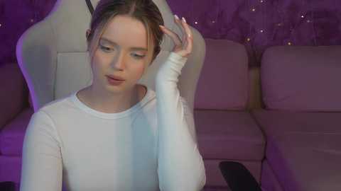 Media: Video of a young woman with light skin and brown hair, wearing a white long-sleeve top, adjusting her hair in a plush gaming chair, against a purple, starry backdrop.