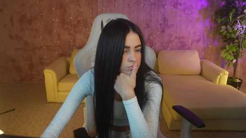 Media: Video of a young woman with long black hair, wearing a light blue sweater, sitting in a beige chair with her hand over her mouth, in a room with a textured brown wall and yellow couch.