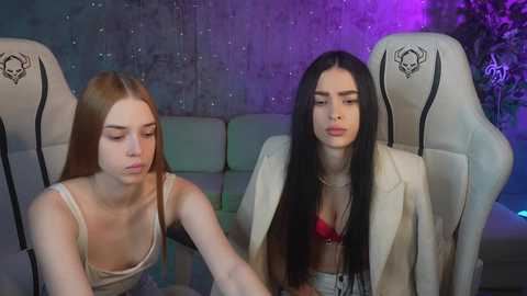 Media: Video of two young women in a modern, dimly lit room. One has long, straight, light brown hair, wearing a tank top, and the other has long, straight, black hair, wearing a white blazer and red bra.