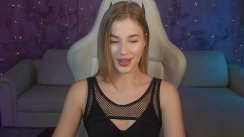 Media: Video of a young Caucasian woman with straight, blonde hair, fair skin, wearing a black, fishnet-patterned dress, seated on a white gaming chair in a dimly lit room with purple, starry wallpaper.